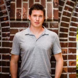 A photo of Andrew Cruickshank wearing a grey shirt, standing in front of a brick wall.