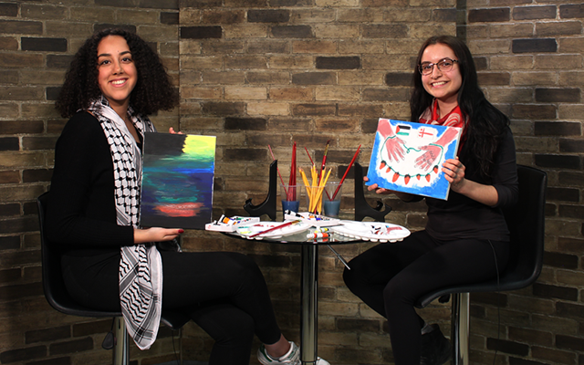 Two people are seated in front of a brick wall, displaying artwork, with paintbrushes in the background.