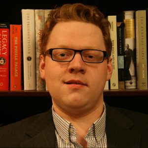 A close up photo of Neil Robert Moss, with books on a shelf visible in the background.