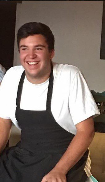 A photo of Jake Tiggelman smiling, wearing a white shirt and black apron.