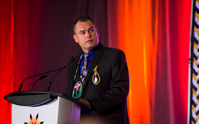 Jesse Thistle, speaking at a podium, with a red curtain in the background.