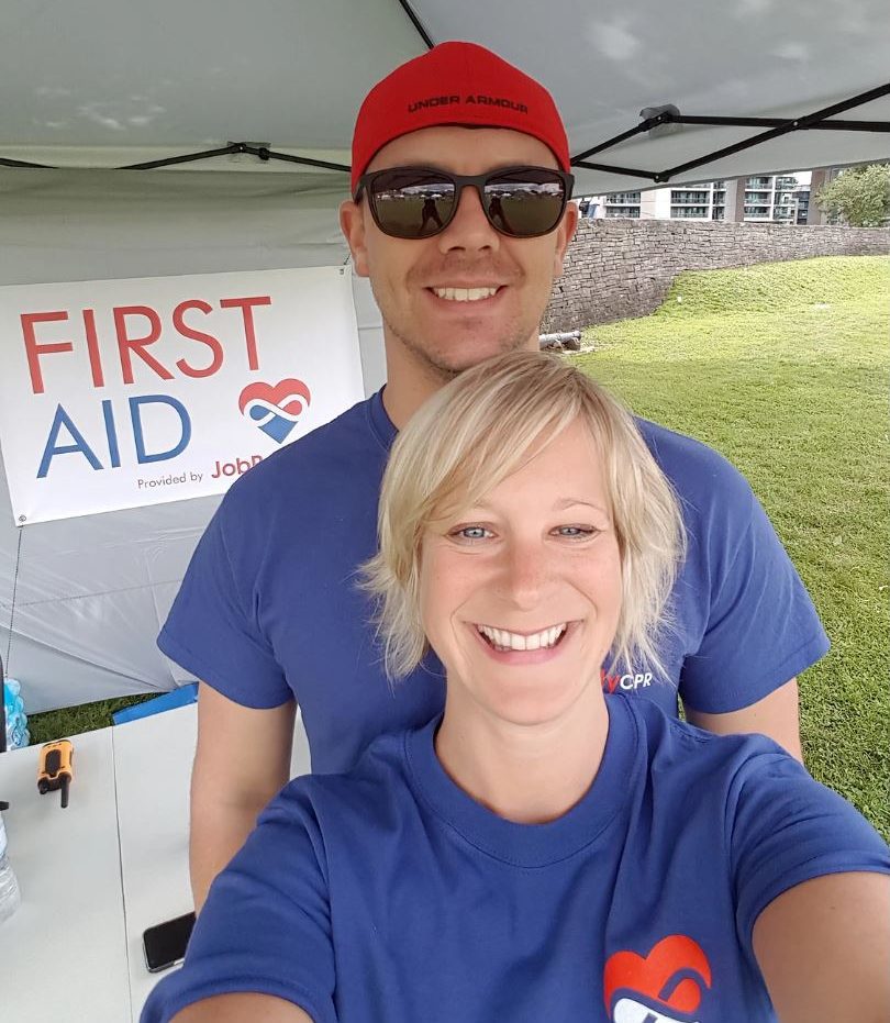 John Budarick is pictured with a paramedical volunteer, with both wearing blue shirts and a sign that says 'FIRST AID' in the background.