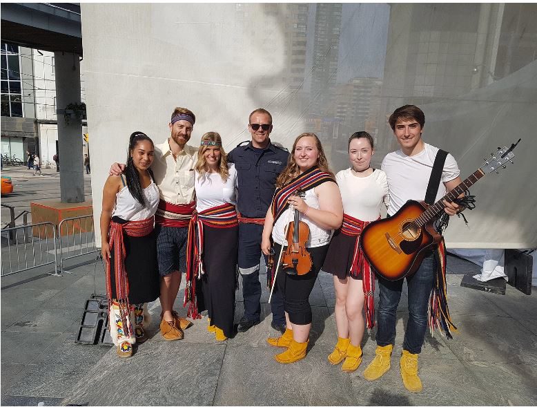 Seven people pose for a photo, with one holding a violin and another holding a guitar.