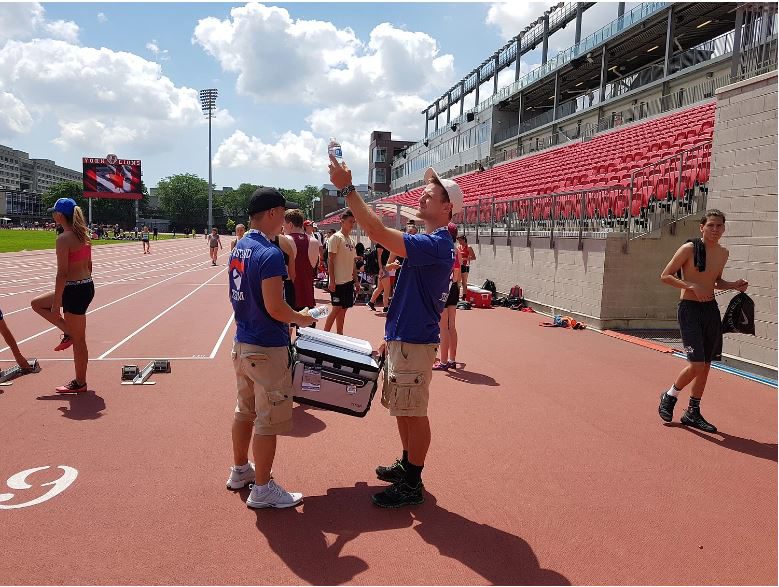 Two people in blue shirts, holding a box on a track, with other people in athletic gear pictured in the background.